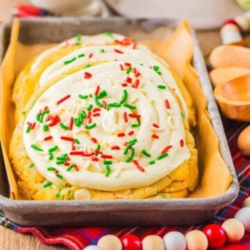 Frosted cookies with red and green sprinkles on a tray, set on a festive plaid cloth with wooden utensils nearby.