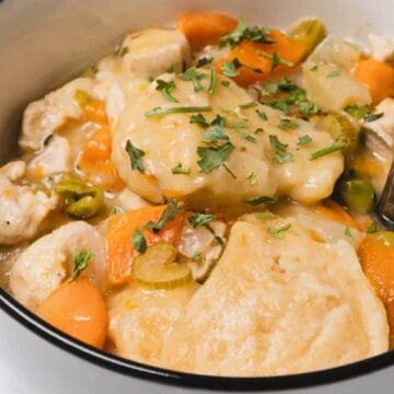 A bowl of chicken and dumplings soup with carrots, celery, and herbs, with a spoon inside.