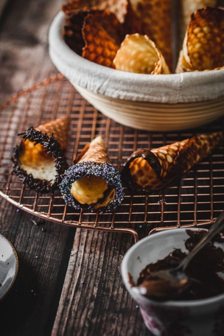 Gluten free ice cream cones with chocolate and sprinkles rest on a wire rack, alongside a bowl of cones and melted chocolate.