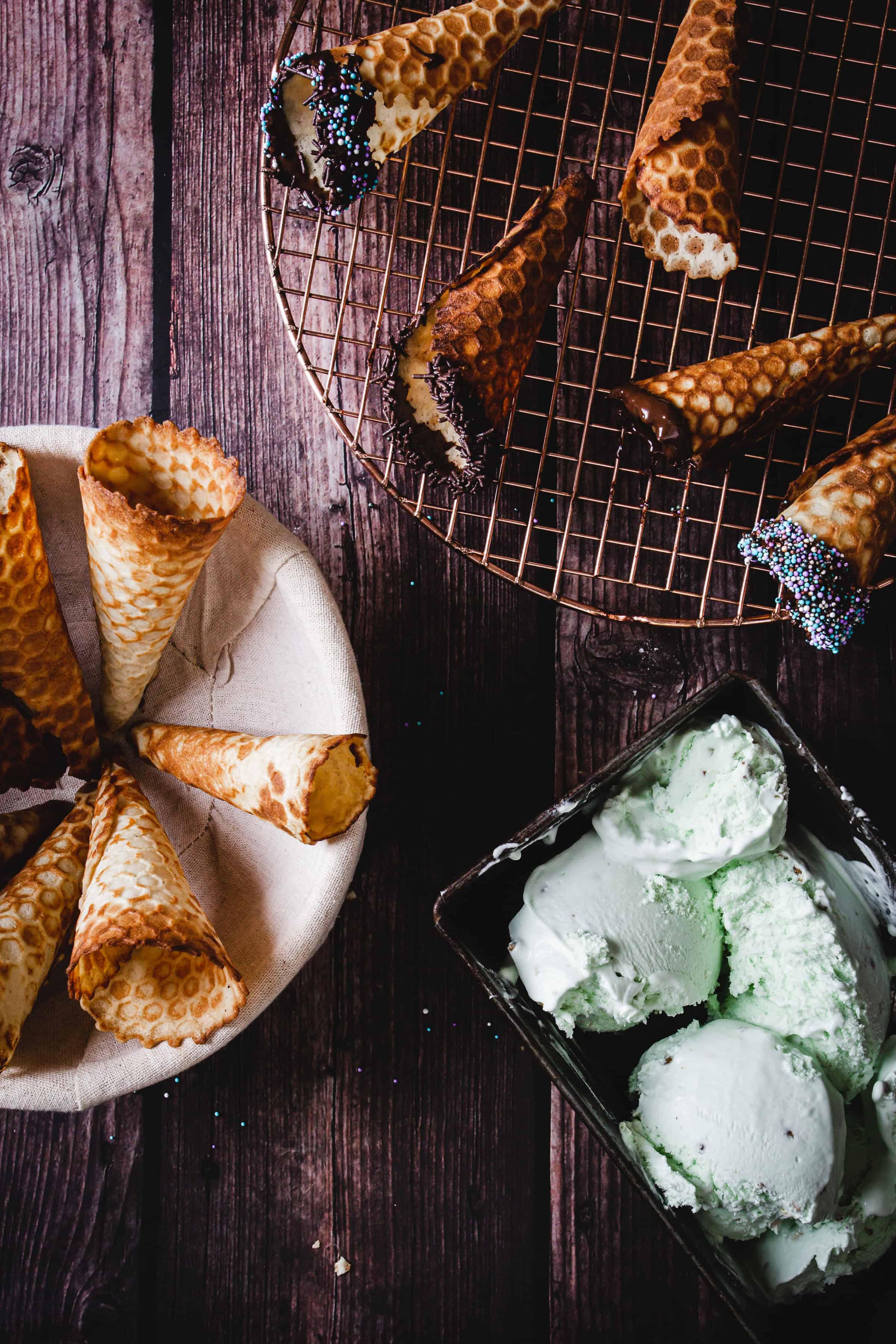 Gluten free ice cream cones and empty waffle cones, some dipped in chocolate and sprinkles, rest on a cooling rack alongside scoops of mint ice cream.