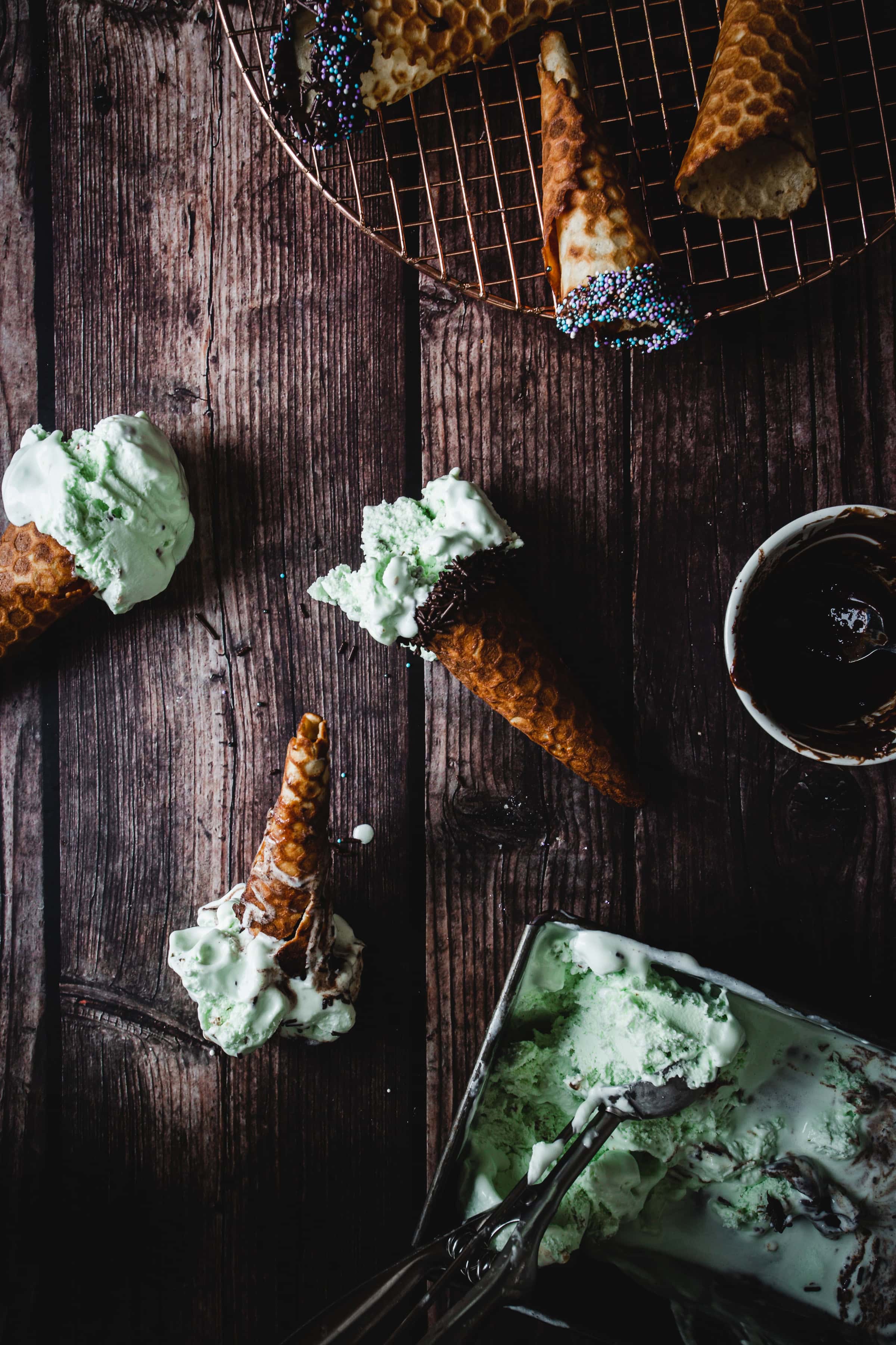 Melted mint ice cream in gluten free ice cream cones on a rustic wooden table, topped with sprinkles and accompanied by a metal ice cream scoop.