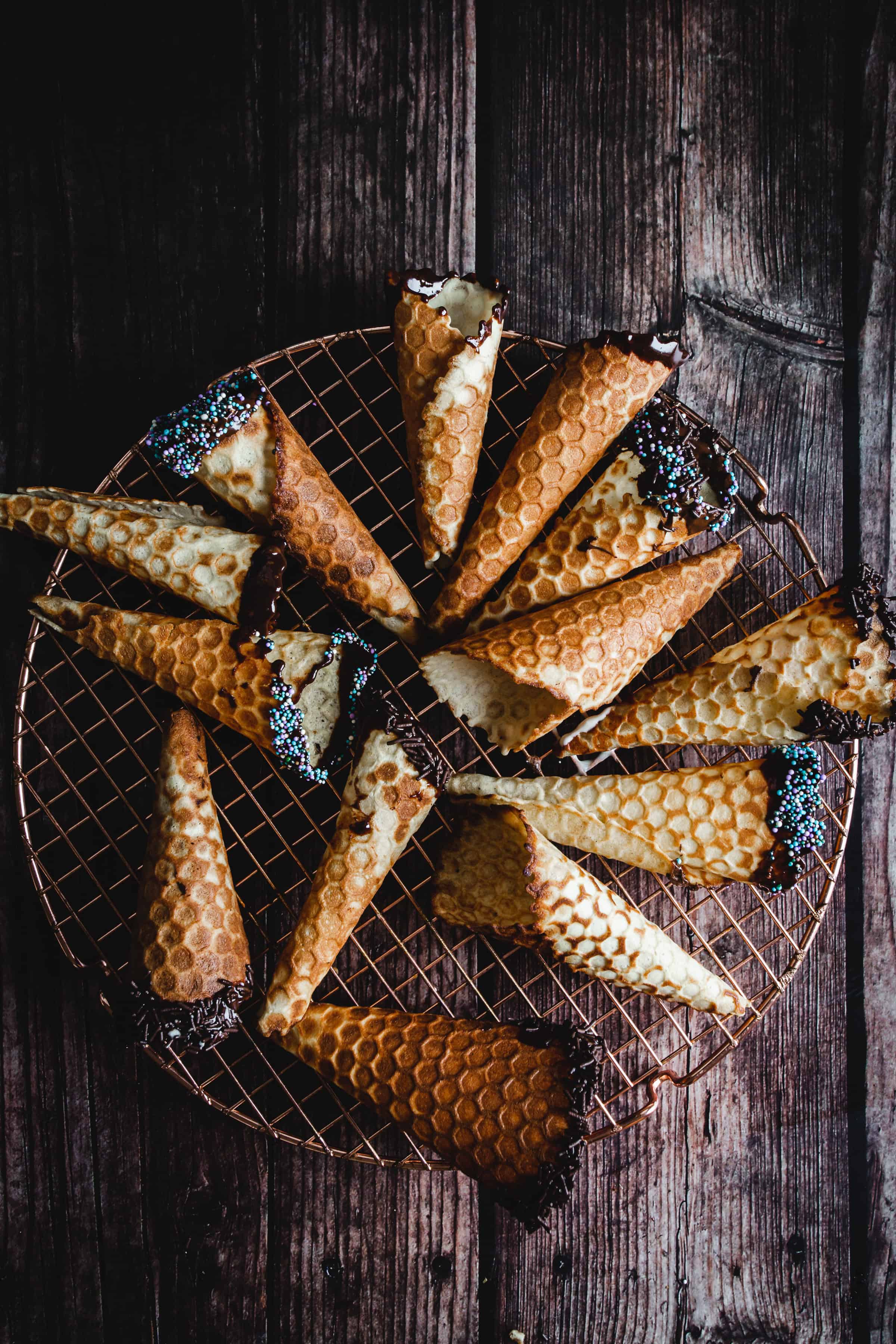 Gluten free ice cream cones dipped in chocolate and sprinkles are arranged on a wire rack over a rustic wooden table.