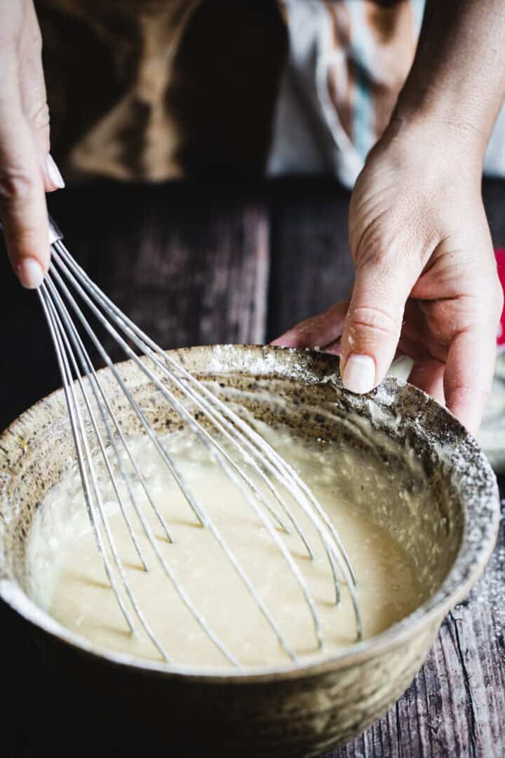 A person whisking batter in a rustic ceramic bowl on a wooden surface, preparing to fill gluten free ice cream cones.