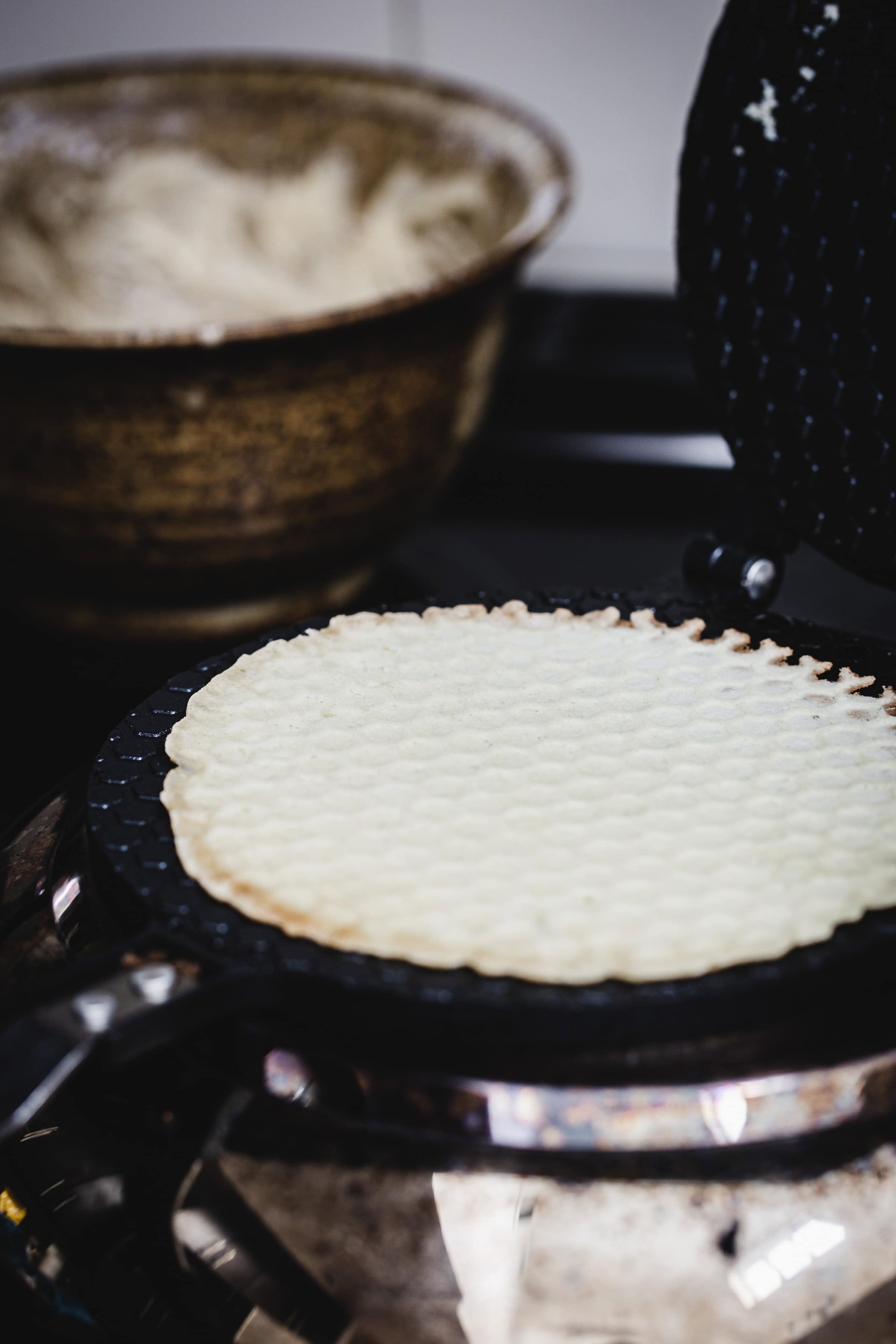 Thin waffle cooking in a waffle iron with a bowl of batter in the background, perfect for making gluten free ice cream cones.