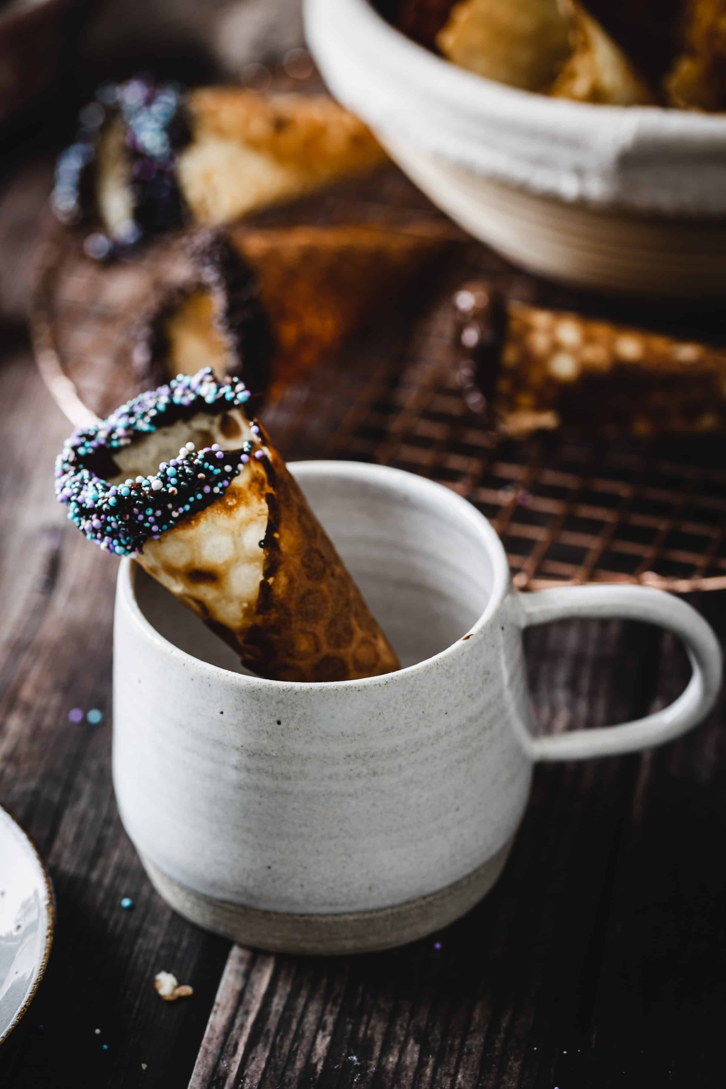 A chocolate-dipped cannoli with sprinkles rests in a white ceramic mug on a wooden table, reminiscent of classic treats like gluten free ice cream cones.