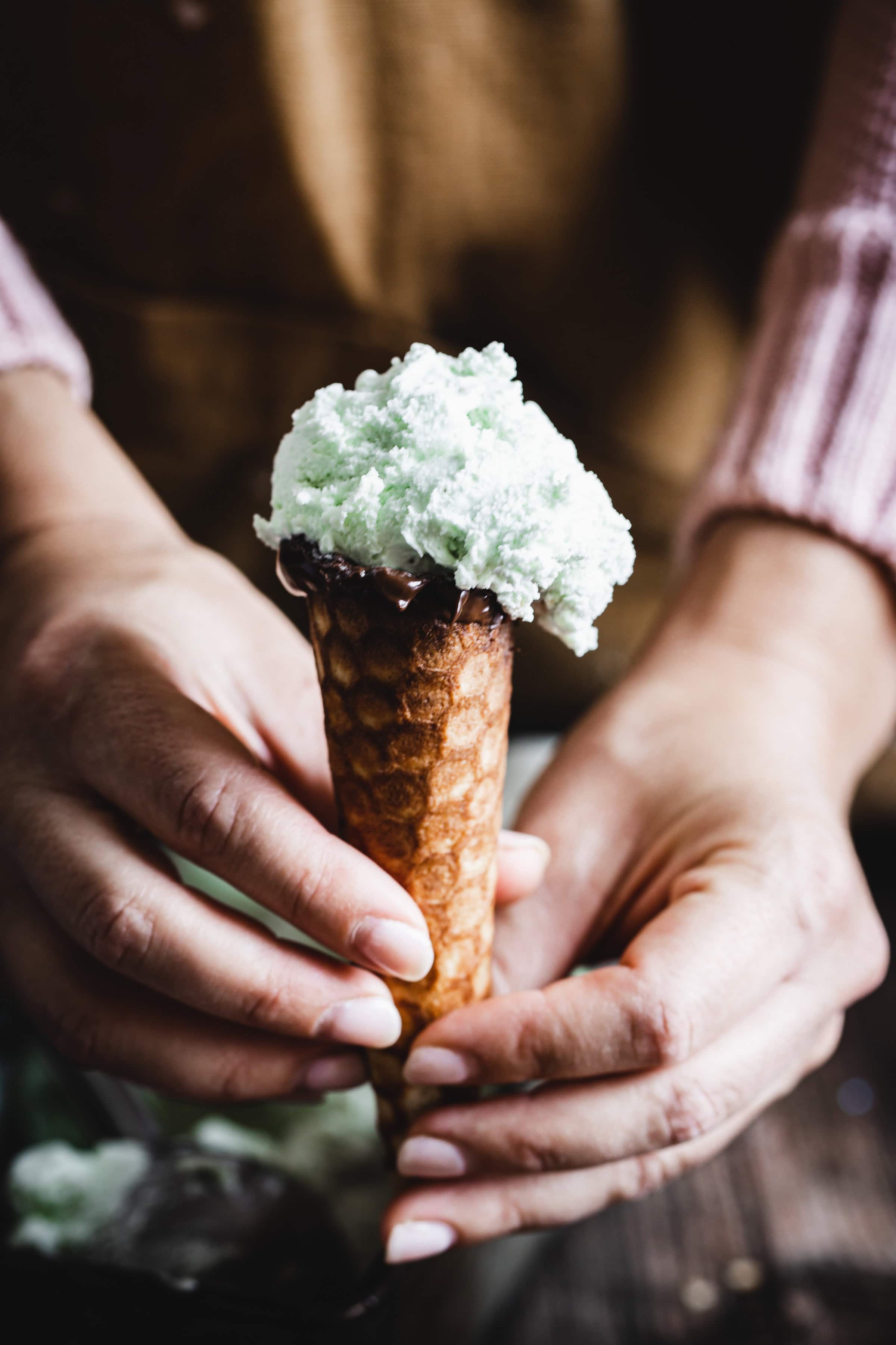 Person holding a gluten free ice cream cone filled with pale green ice cream, wearing a brown apron and pink sweater.