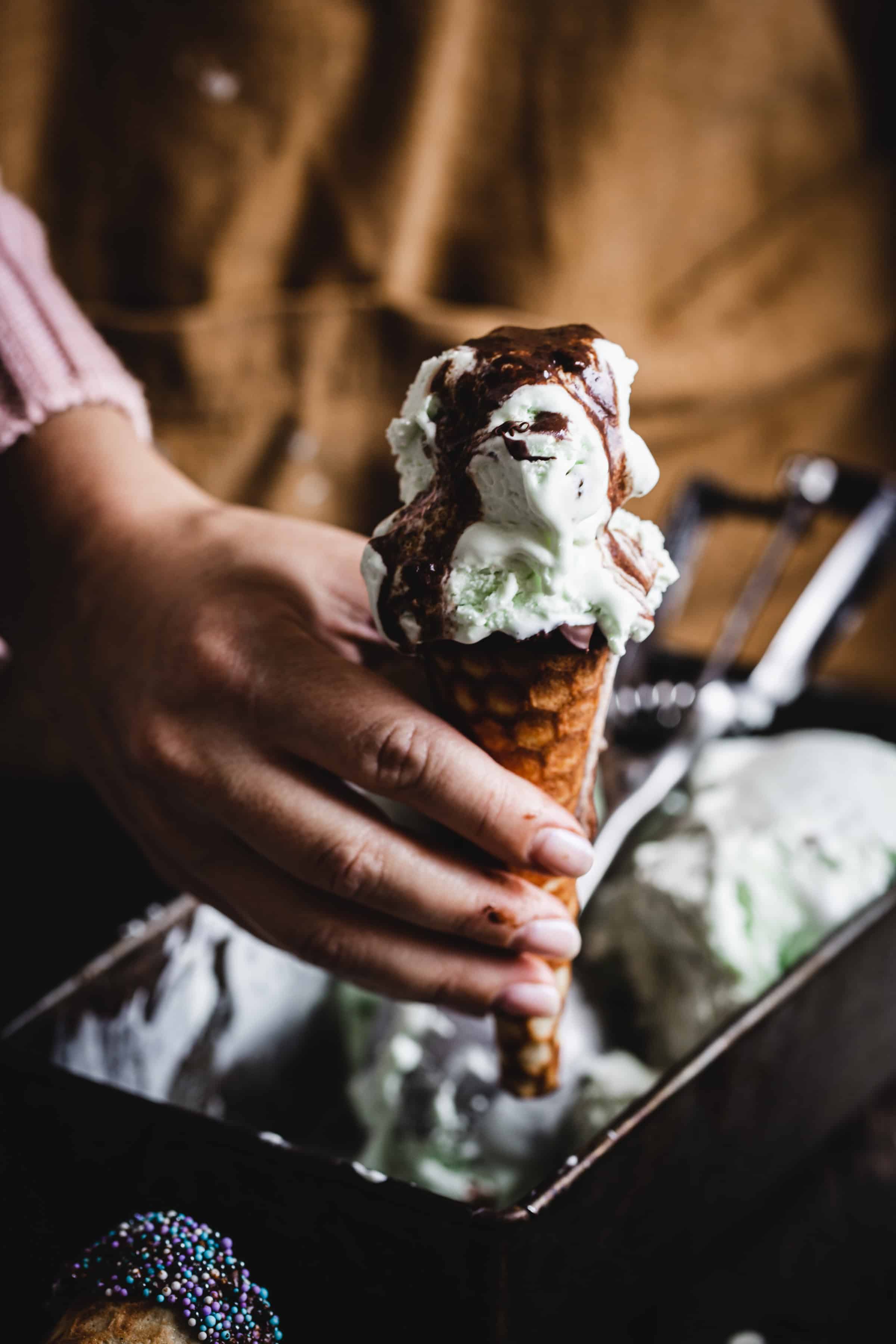A hand holding a gluten free ice cream cone filled with mint ice cream and chocolate syrup, poised over a container of ice cream.