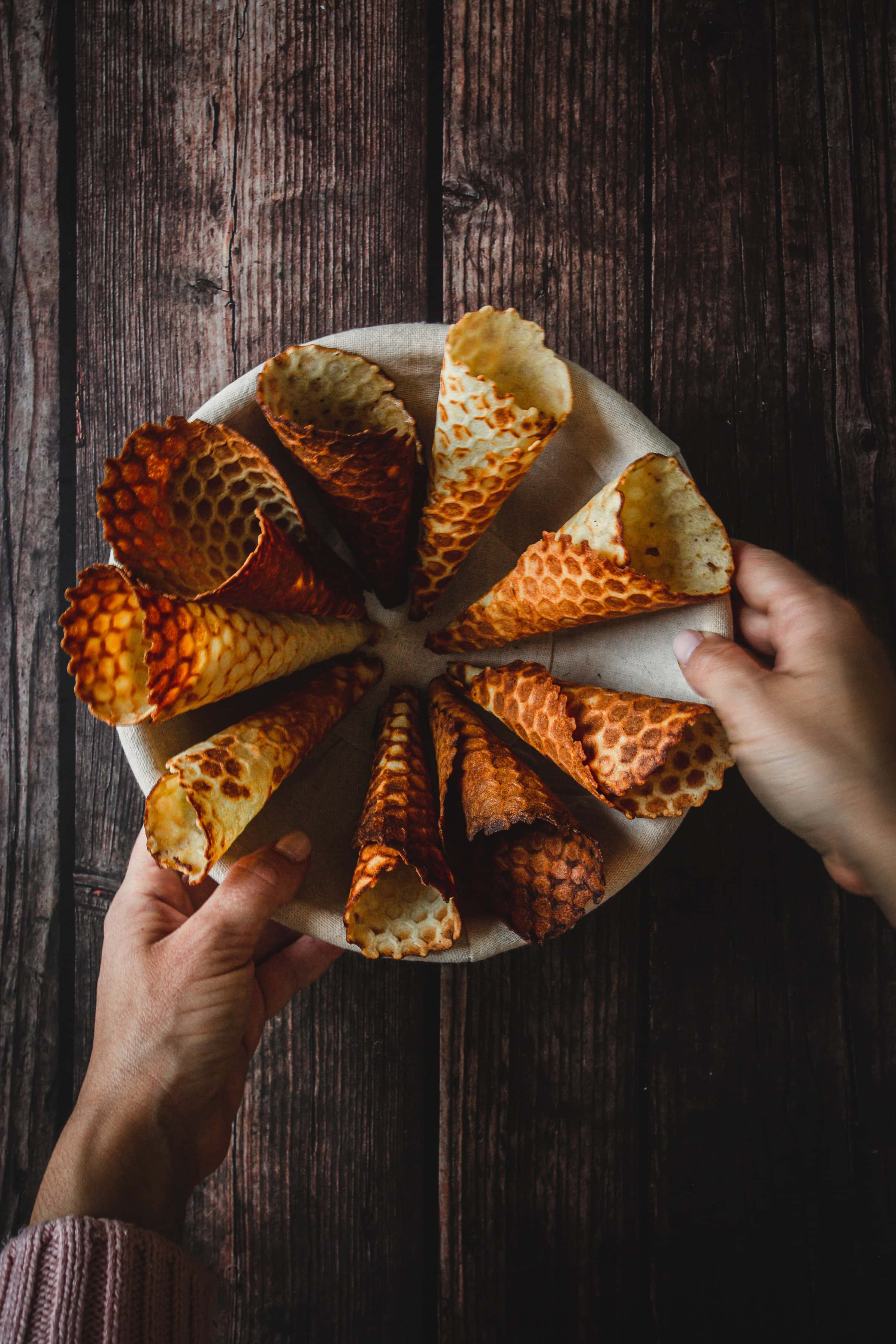 Hands holding a plate with a circular arrangement of gluten free ice cream cones on a dark wooden surface.