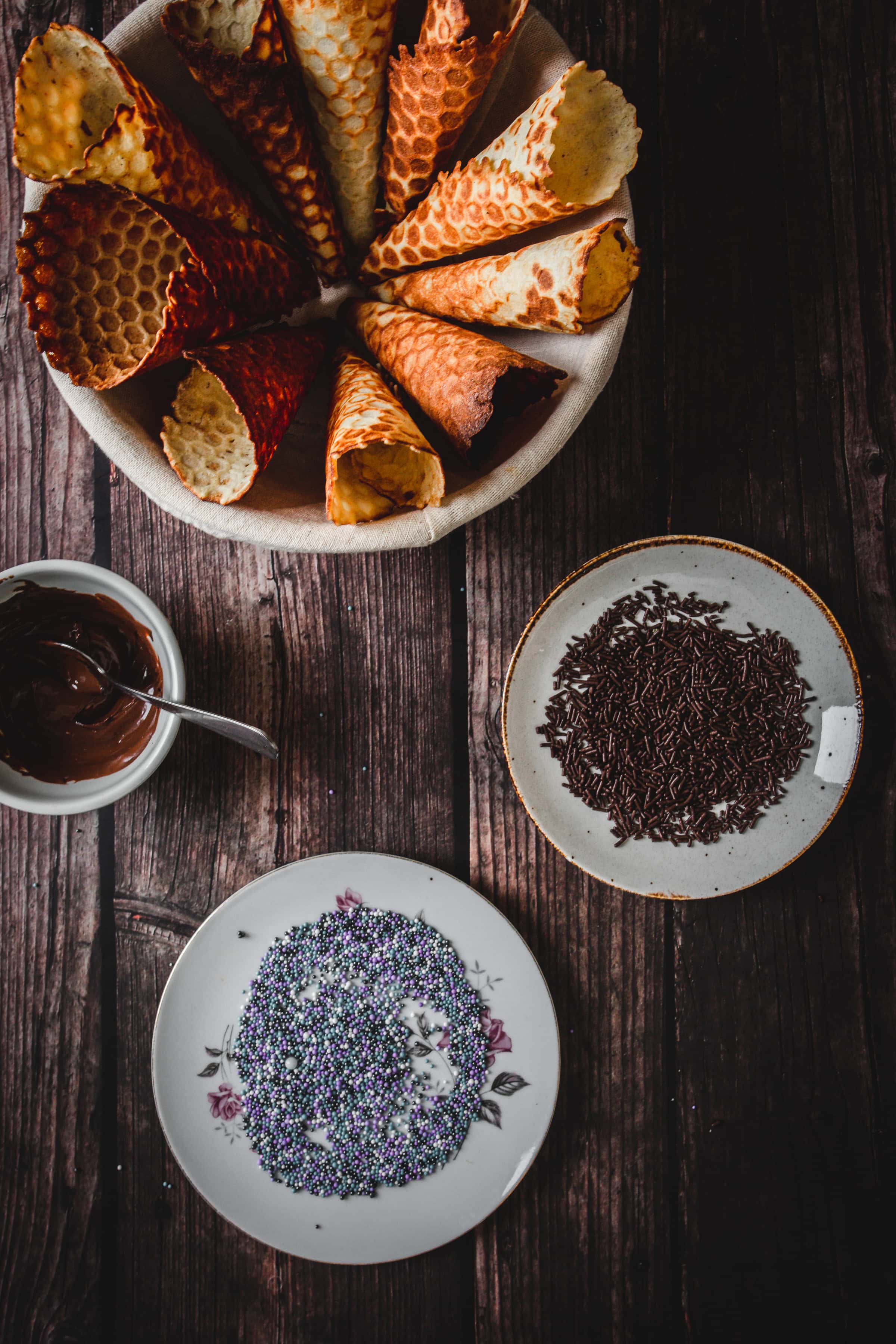A bowl of gluten free ice cream cones, a plate of chocolate sprinkles, and a plate of colorful sprinkles on a wooden table.