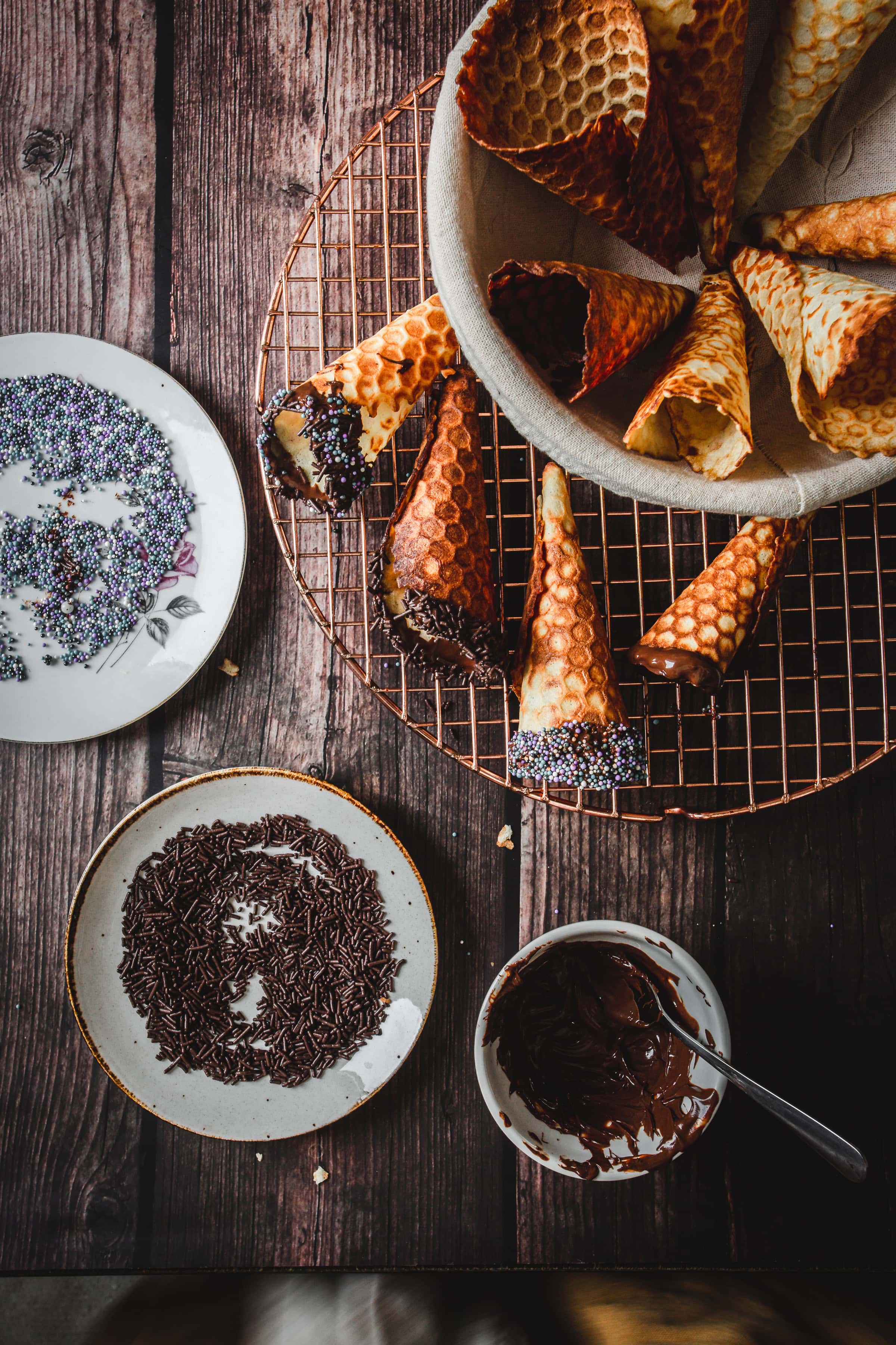 Gluten free ice cream cones dipped in chocolate and sprinkles rest on a cooling rack, alongside plates filled with colorful sprinkles and melted chocolate.