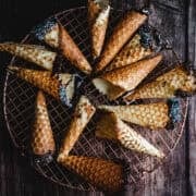 Gluten free ice cream cones with chocolate and sprinkles rest on a cooling rack over a rustic wooden surface.