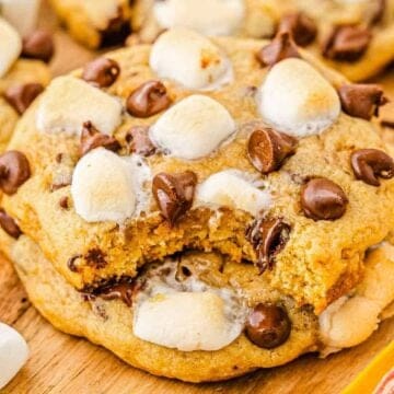 A chocolate chip cookie with marshmallows, missing a bite, on a wooden board with more cookies and marshmallows.