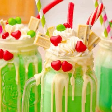 Three green milkshakes in mason jars, topped with whipped cream, candy, and white chocolate pieces.