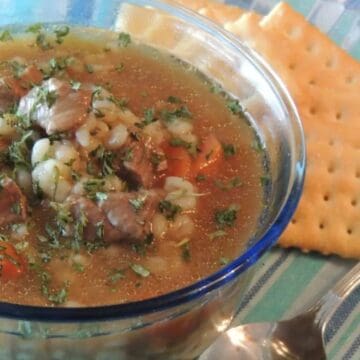 A bowl of beef and barley soup with herbs, served with saltine crackers on a plate and a spoon.