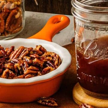 Small orange dish of candied pecans next to a mason jar filled with dark syrup on a table.