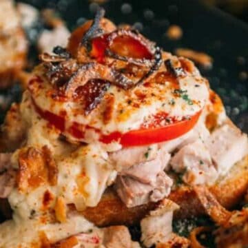 Toasted bread topped with turkey, sliced tomato, creamy sauce, bacon, and melted cheese, garnished with herbs.