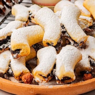 A plate of powdered sugar-dusted filled cookies sits on a table with nuts and holiday decor in the background.