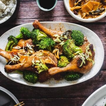 Oval plate of roasted chicken legs with broccoli and scallions, surrounded by rice and side dishes on a wooden table.