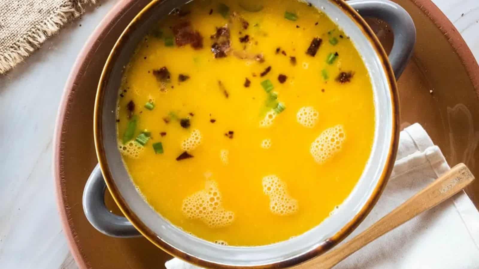 A bowl of creamy yellow soup garnished with herbs and bits of crispy bacon, with a wooden spoon beside it.