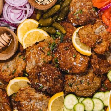 A platter with latkes, lemon slices, cucumbers, red onions, pickles, carrots, dip, and honey.