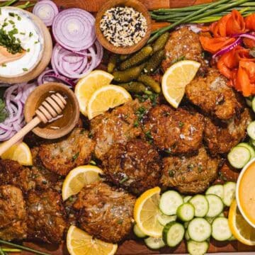 A platter with latkes, lemon slices, cucumber, onions, smoked salmon, sauces, herbs, and pickles.