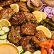 Crispy potato pancakes with lemon slices, cucumber, red onion, pickles, and honey on a platter.