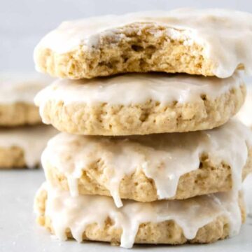Three thick, iced cookies stacked, with one bitten, and more cookies blurred in the background.