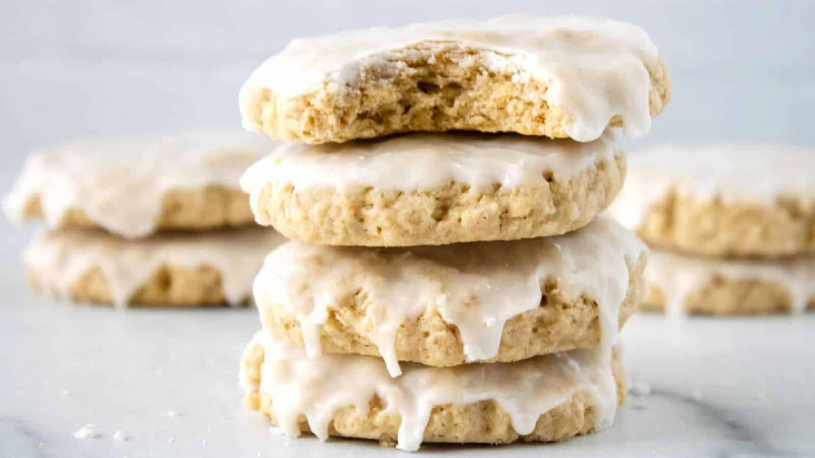 Three thick, iced cookies stacked, with one bitten, and more cookies blurred in the background.