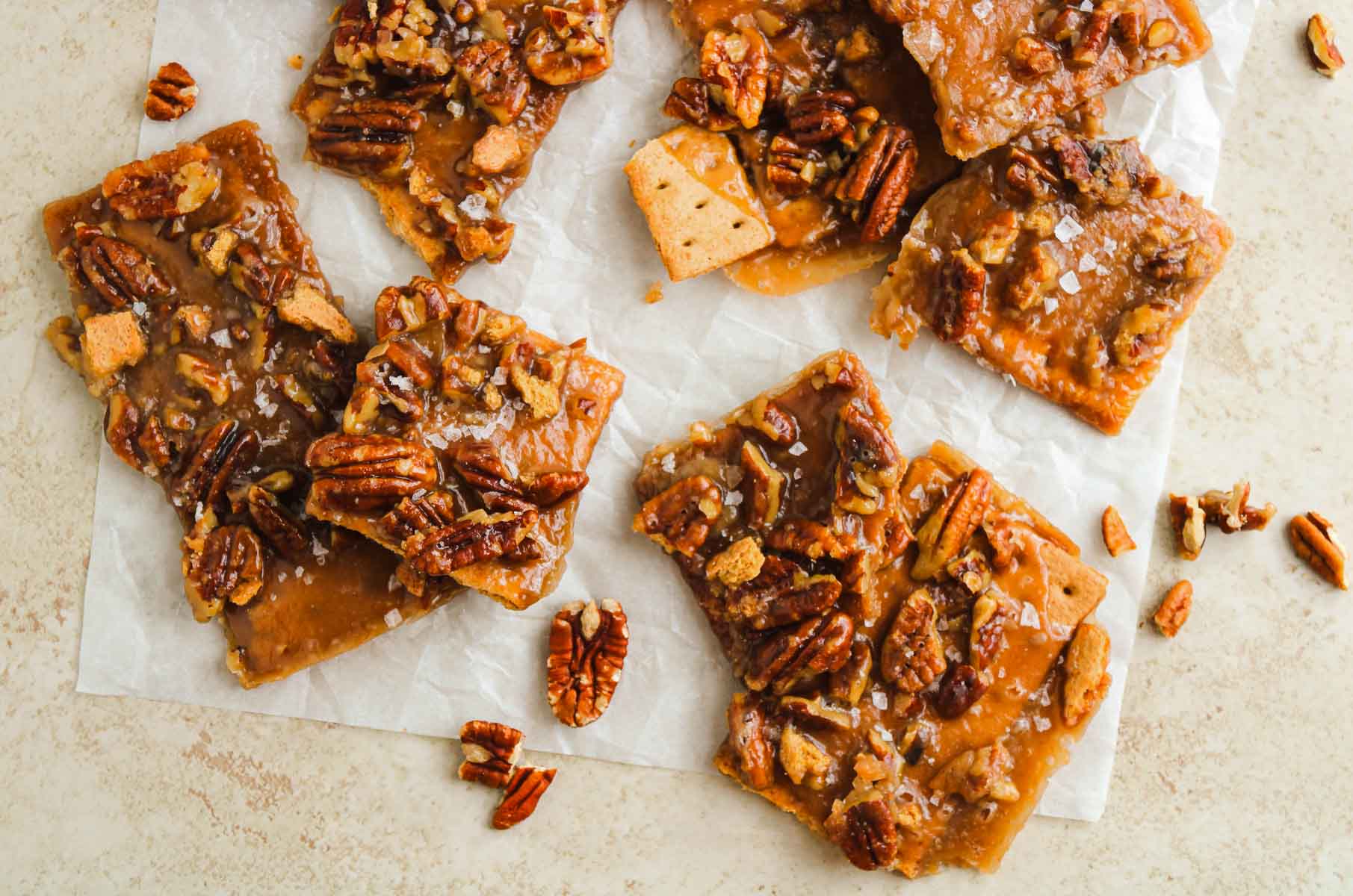 Caramel pecan bars with graham cracker base on parchment paper, sprinkled with sea salt and pecan pieces.