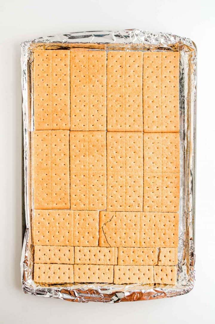 A baking sheet lined with foil holds neatly arranged graham crackers, some of which are cracked.