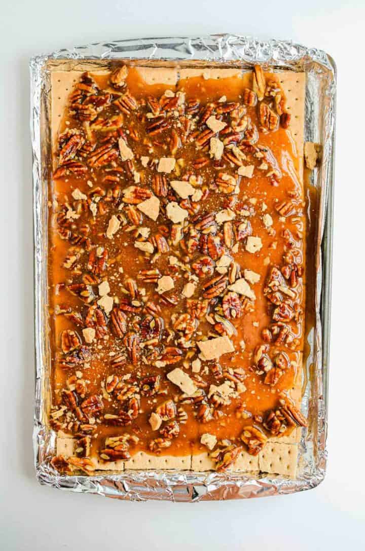 A foil-lined tray with crackers topped with caramel, pecans, and crushed crackers.