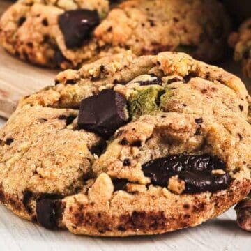 A close-up of a chunky cookie with chocolate pieces and pistachios on a wooden board.