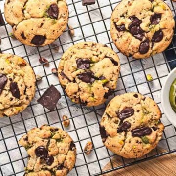 Chocolate chunk cookies with nuts on a cooling rack, next to a bowl of green spread and chocolate pieces.
