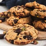 A pile of chunky chocolate chip cookies with pistachios on a wooden board.