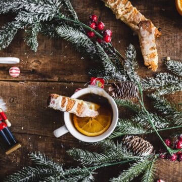 Mugs with biscotti, orange slices, pine branches, and Christmas decorations on a rustic wooden table.