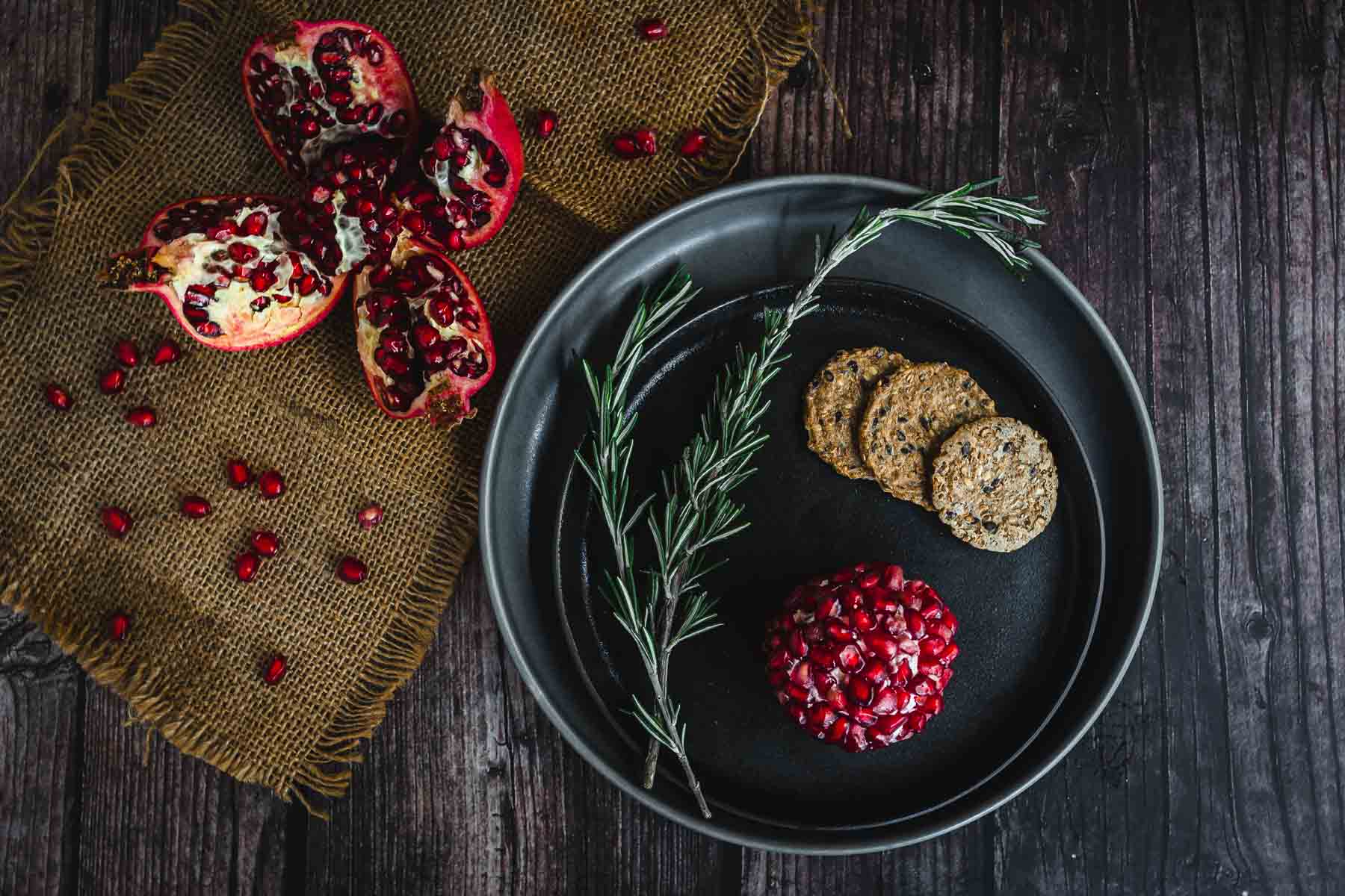 Black plate with crackers, rosemary, and a festive Christmas Cheese Ball coated in pomegranate seeds, next to halved pomegranates.