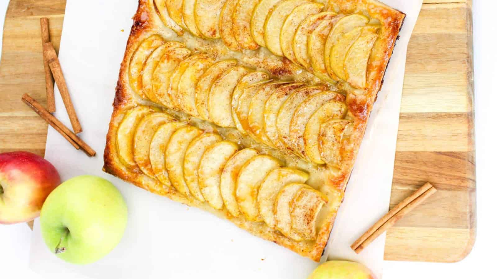 Rectangular apple tart with golden apple slices, a flaky crust, and a light glaze. Two cinnamon sticks lie nearby on a wooden surface.