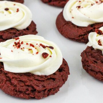 Red velvet cookies topped with swirls of white frosting and small red crumbs on a white plate.