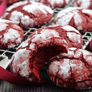 Red velvet crinkle cookies with powdered sugar, one with a bite, on a cooling rack with a red ribbon—perfect for gifting or as easy Christmas cookies.