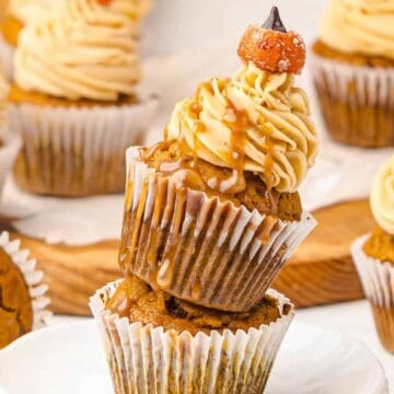 Pumpkin cupcakes with swirled frosting and candy toppers, stacked on a plate with more cupcakes in the background.