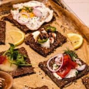 Crispy flatbread squares topped with eggs, veggies, herbs, and lemon wedges on a parchment-lined tray.