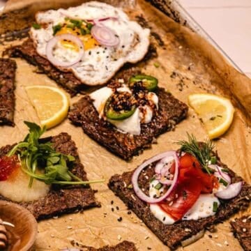 Crispy flatbread squares topped with eggs, veggies, herbs, and lemon wedges on a parchment-lined tray.