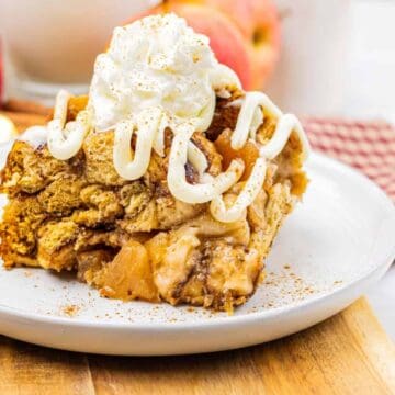A slice of apple pie with whipped cream on top, served on a white plate with apples in the background.