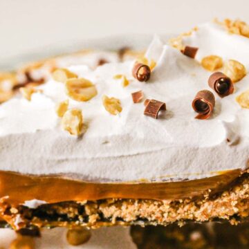 A close-up of a creamy pie slice with whipped topping, chocolate curls, and chopped nuts in a crumb crust.