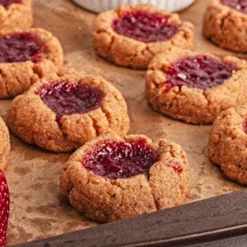 Thumbprint cookies with strawberry jam on a baking tray, surrounded by fresh strawberries.