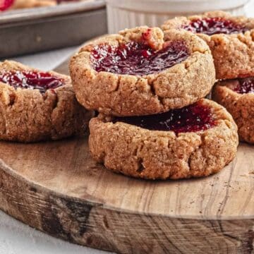 Stack of thumbprint cookies with red jam filling on a wooden board, strawberries in the background.