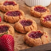 Close-up of thumbprint cookies with red jam filling on parchment paper, with fresh strawberries nearby.