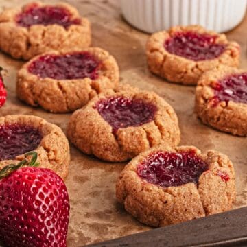 Close-up of thumbprint cookies with red jam filling on parchment paper, with fresh strawberries nearby.