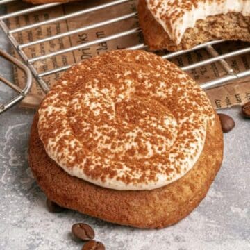 A frosted cookie dusted with cocoa powder, surrounded by coffee beans and baked cookies on a cooling rack.