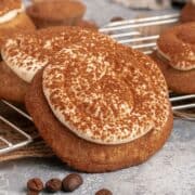 A cookie topped with cream and dusted with cocoa powder, next to coffee beans and more cookies on a rack.